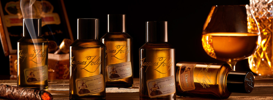 Fünf Jacques Zolty Parfumflakons stehen auf einem rustikalen Holztisch, daneben ein brennender Zigarrenstummel und ein Glas Cognac – im Hintergrund Zigarrenkiste und Karaffe.
