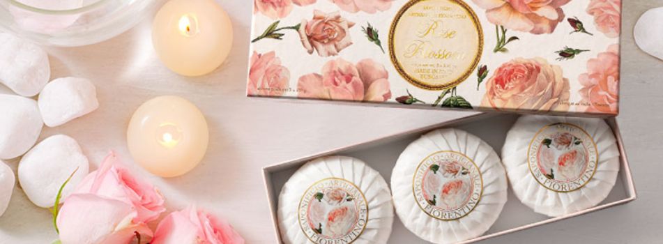 Saponificio Artigianale Fiorentino Geschenkset mit drei runden Rosenseifen in eleganter Verpackung mit Rosendekor, umgeben von Rosenblüten, weißen Steinen und brennenden Kerzen.