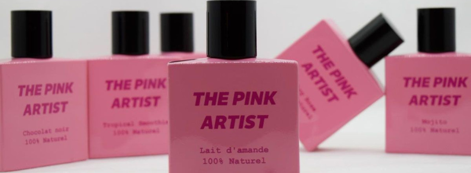 Mehrere pinkfarbene Parfumflakons der Marke ‚The Pink Artist‘ mit schwarzem Verschluss. Auf den Flakons stehen verschiedene Duftnamen wie ‚Lait d’amande‘, ‚Chocolat noir‘ und ‚Mojito‘, jeweils mit dem Hinweis ‚100% Naturel‘.