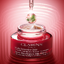 Clarins Multi-Intensive Jour Tagescreme in einem roten Glastiegel mit weißem Deckel. Über dem Tiegel befindet sich ein gold-grüner Tropfen in einer Pipette. Der Hintergrund ist in einem kräftigen Rotton mit Lichtkreisen gestaltet.
