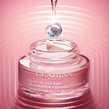 Clarins Multi-Active Jour Tagescreme in einem rosafarbenen Glastiegel mit weißem Deckel. Ein rosa-violetter Tropfen mit Pflanzenmotiv hängt über dem Tiegel. Der Hintergrund zeigt sanfte rosa Lichtwellen.