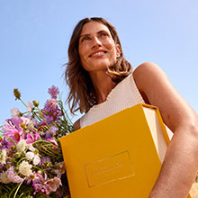 Lächelnde Frau im Sonnenlicht mit einem gelben L'OCCITANE Geschenkkarton und Blumenstrauß.