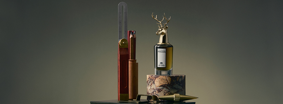 Penhaligon’s Parfum 'The Tragedy of Lord George' aus der Portraits-Kollektion. Der markante Flakon mit goldener Hirschkopf-Kappe steht neben Maßwerkzeugen auf einem Marmorsockel.