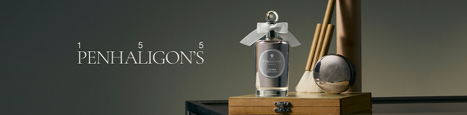 Penhaligon’s Parfumflakon 'Luna' steht auf einer Holzbox, umgeben von feinen Kosmetikpinseln und einer Puderdose. Der Flakon ist elegant mit einer weißen Schleife verziert. Links im Bild steht in stilvoller Schrift: 'Penhaligon’s 155'.