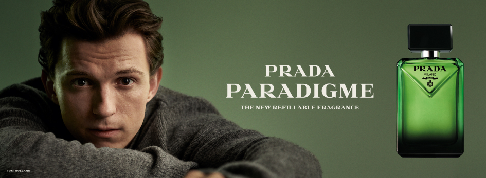 Werbebanner für den Herrenduft Prada Paradigme: Links ein Mann in grauem Pullover, der den Betrachter direkt ansieht, rechts eine grün-schwarze Parfumflasche mit schwarzem Verschluss und dem Schriftzug ‚Prada Milano‘. In der Mitte steht der Text ‚Prada Paradigme – The new refillable fragrance‘.
