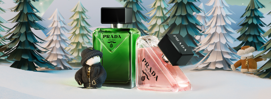 Zwei Parfümflakons von Prada, einer in Grün und einer in Rosa, stehen in einer winterlichen Schneelandschaft mit Tannenbäumen und kleinen Stofffiguren. Die Szene vermittelt ein festliches, weihnachtliches Ambiente.
