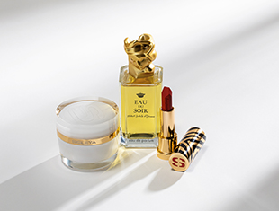 Drei luxuriöse Kosmetikprodukte auf heller Fläche mit weichem Schatten: ein gläserner Tiegel mit weißer Creme und goldener Verzierung, ein Parfümflakon mit goldener Verschlusskappe und gelbem Etikett mit der Aufschrift 'Eau du Soir', sowie ein geöffneter goldener Lippenstift mit roter Farbe und einem Deckel mit schwarzem Muster und 'S'-Logo.