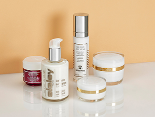 Fünf Pflegeprodukte von Sisley Paris auf weißer Oberfläche vor beigem Hintergrund: eine rote Cremedose, ein Pumpspender mit durchsichtigem Behälter und silbernem Deckel, eine weiße Flasche mit silbernem Spender, sowie zwei gläserne Cremetiegel mit weißem Inhalt und goldenen Zierstreifen.