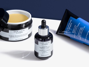 Drei Haarpflegeprodukte der Linie 'Hair Rituel by Sisley' stehen auf einer hellen Fläche vor einem dunkelblauen Hintergrund. Links ist ein offener Tiegel mit gelblicher Pflegecreme zu sehen, in der Mitte eine kleine Pipettenflasche mit Serum, rechts eine liegende Tube mit blau-schwarzem Design. Die Produkte wirken hochwertig und sind für Haar- und Kopfhautpflege konzipiert.