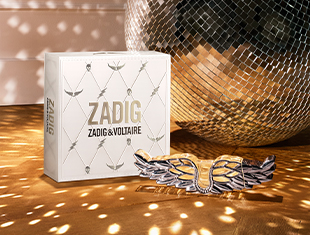 Weißes Geschenkset von Zadig & Voltaire mit Flügelornament aus Metall im Vordergrund. Die Box ist mit dezenten Linien und kleinen Engelsflügeln verziert, im Hintergrund reflektiert eine große silberne Diskokugel das Licht.